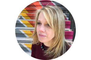 A profile photo of a white woman with shoulder-length blonde hair. She sits on a flight of steps colourfully painted in an ascending chevron pattern.