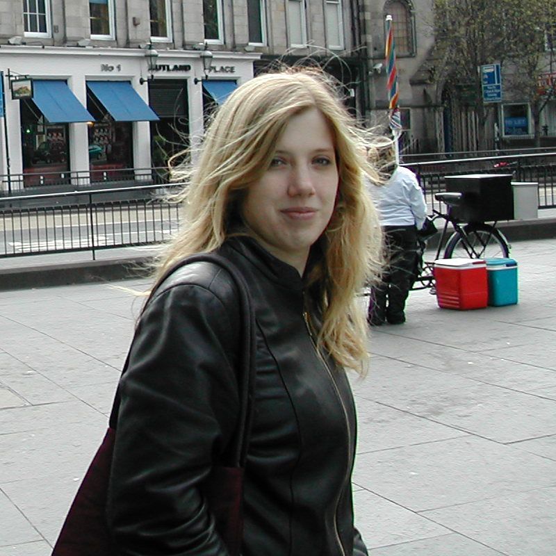 twenty-something white woman with long blonde hair and wearing a black leather jacket, semi-smiles to camera as she’s walking along the street, slightly caught off guard by the unexpected photo.